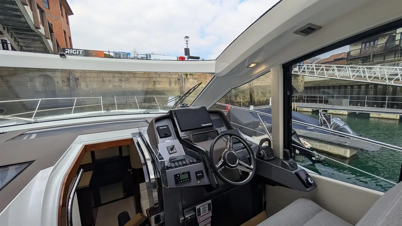 Boat cockpit view featuring a steering wheel, navigation controls, and a display screen, overlooking a marina with boats.